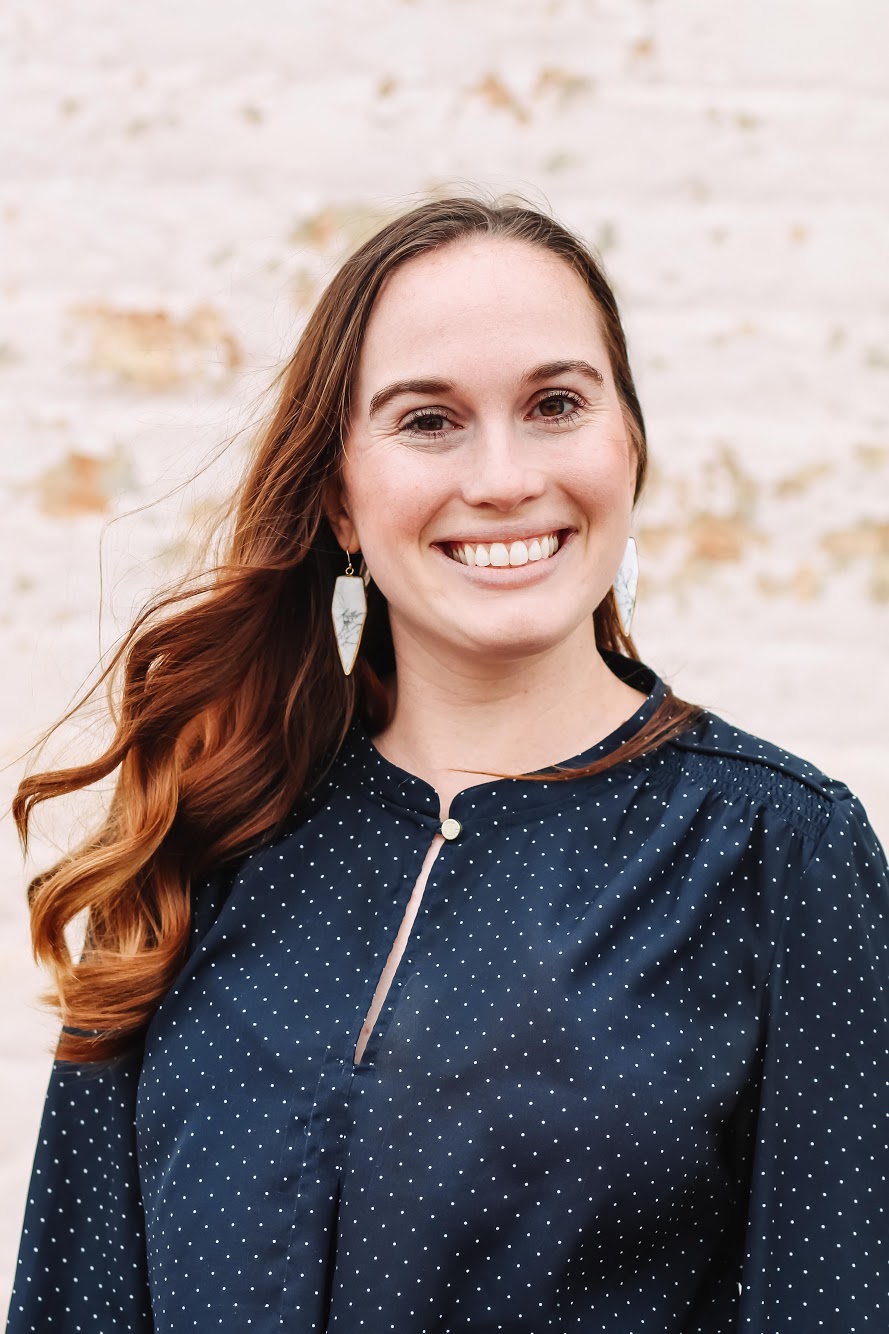 Smiling woman with long brown hair wearing a navy blue polka dot blouse, standing outdoors with wind gently blowing her hair