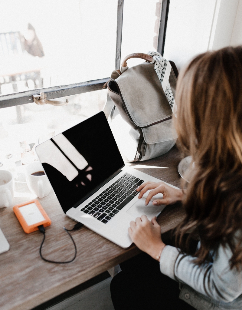 Brunette woman working on a laptop in a bright, modern workspace, focusing on her screen with natural light coming in through large windows.