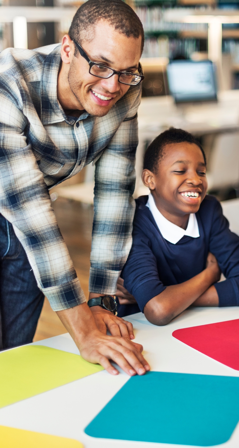 Student and teacher smiling and working together at a desk in a classroom, focusing on a project with books and papers spread out in front of them.