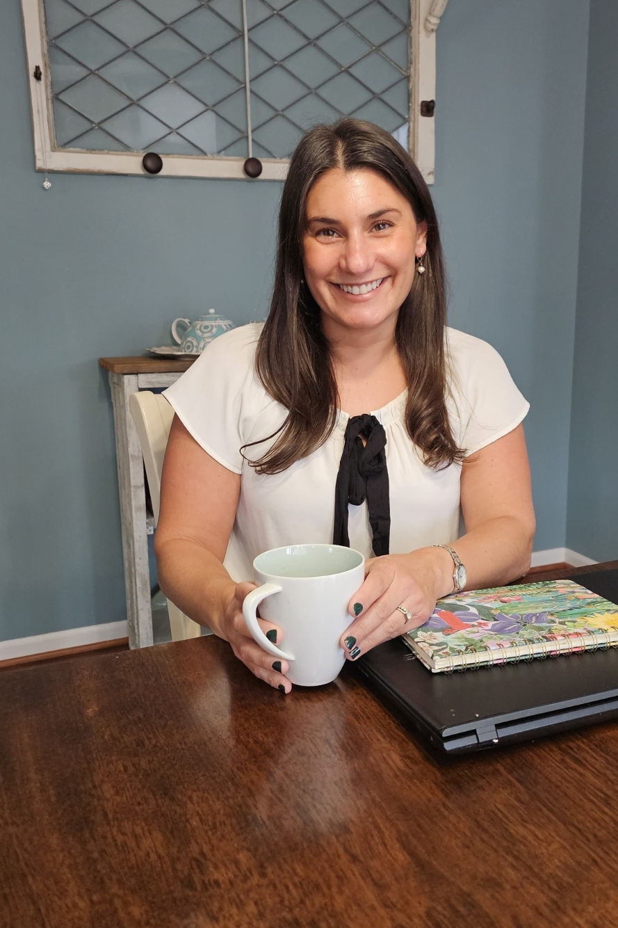 A smiling brunette woman sitting at a table with a coffee mug and work supplies.